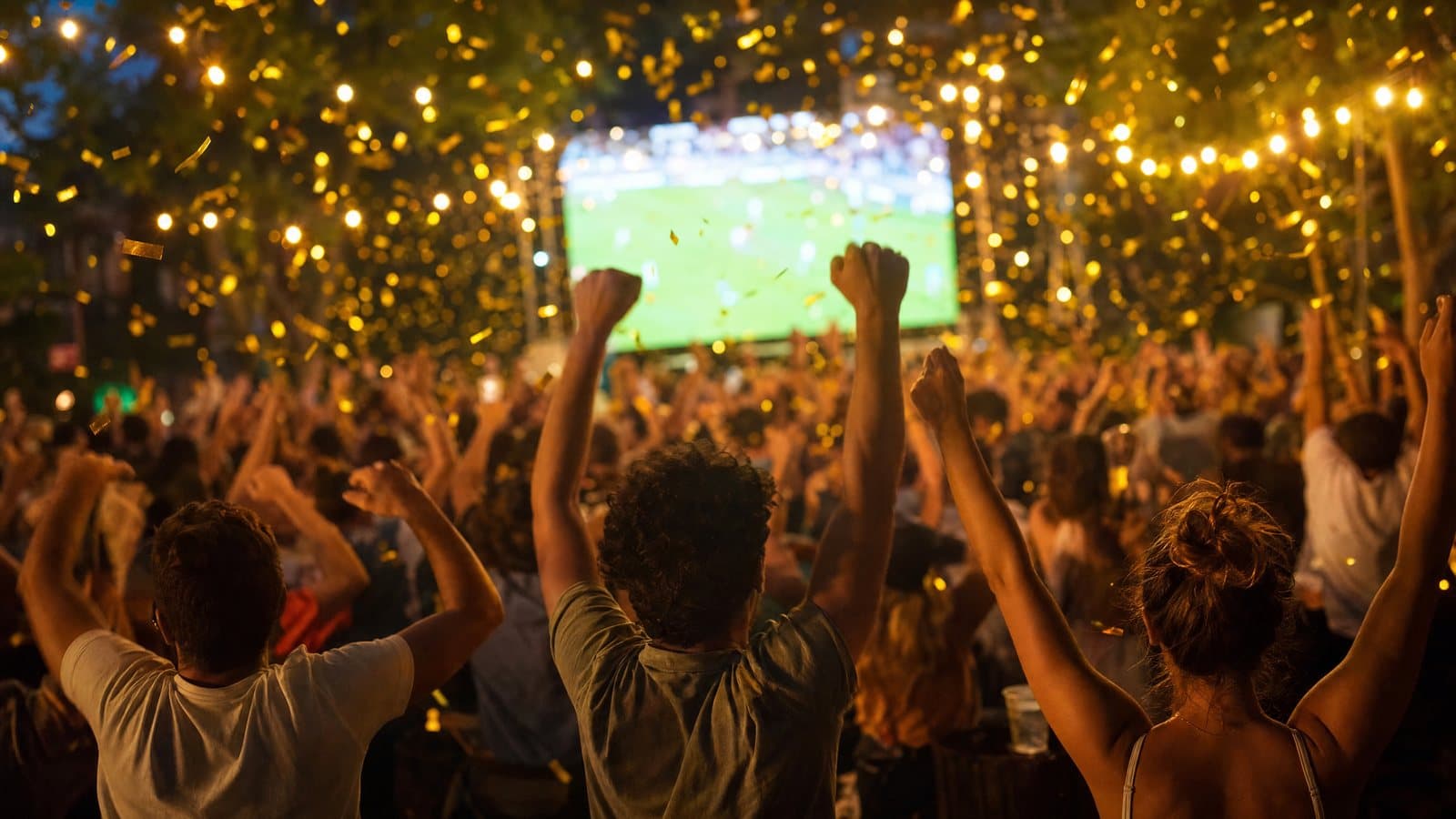 Symbolbild: Jubelnde Menschenmenge von hinten vor einer Leinwand mit einem Fußballspiel, erhobene Fäuste, Lichterketten und Goldkonfetti.